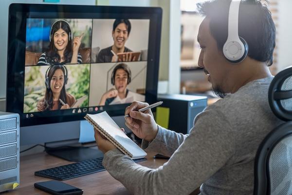 A student wearing headphones sits in front of a computer monitor showing four smiling virtual meeting participants.